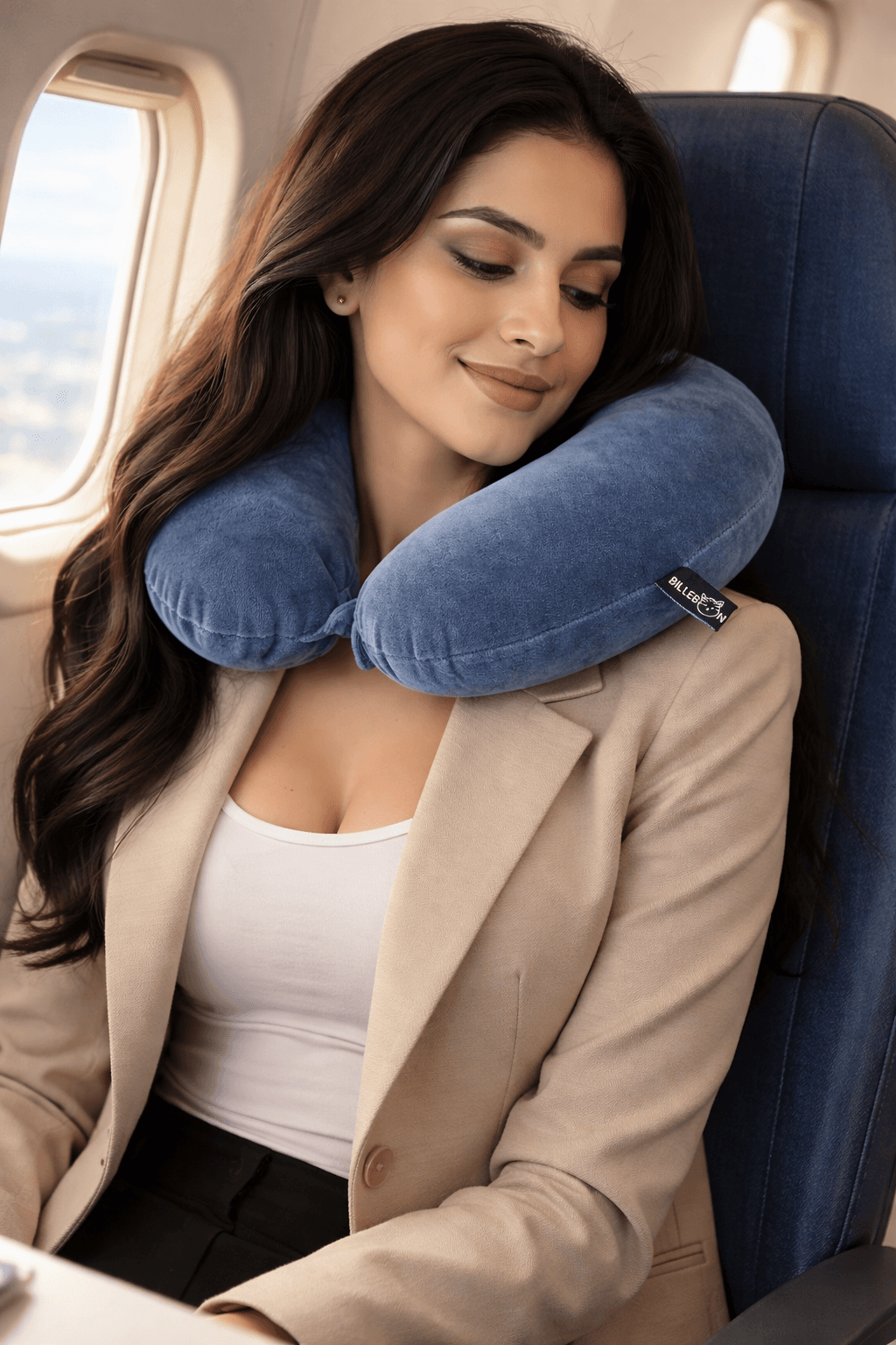 Woman in a beige blazer rests comfortably in an airplane seat using a blue neck pillow.